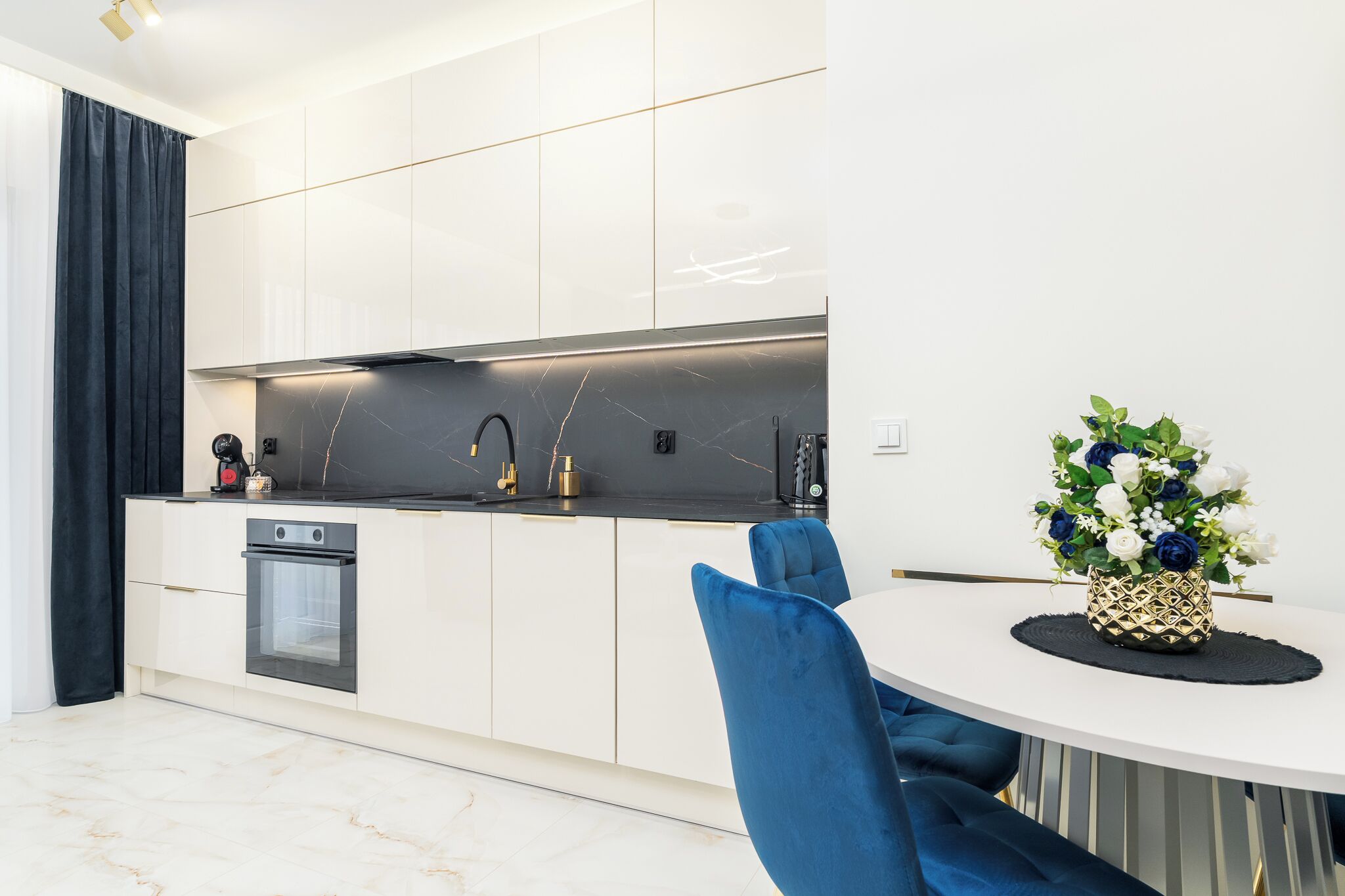 Una cucina moderna con mobili bianchi, un piano di lavoro nero e un forno integrato. C'è un tavolo da pranzo con sedie blu e un vaso di fiori.
