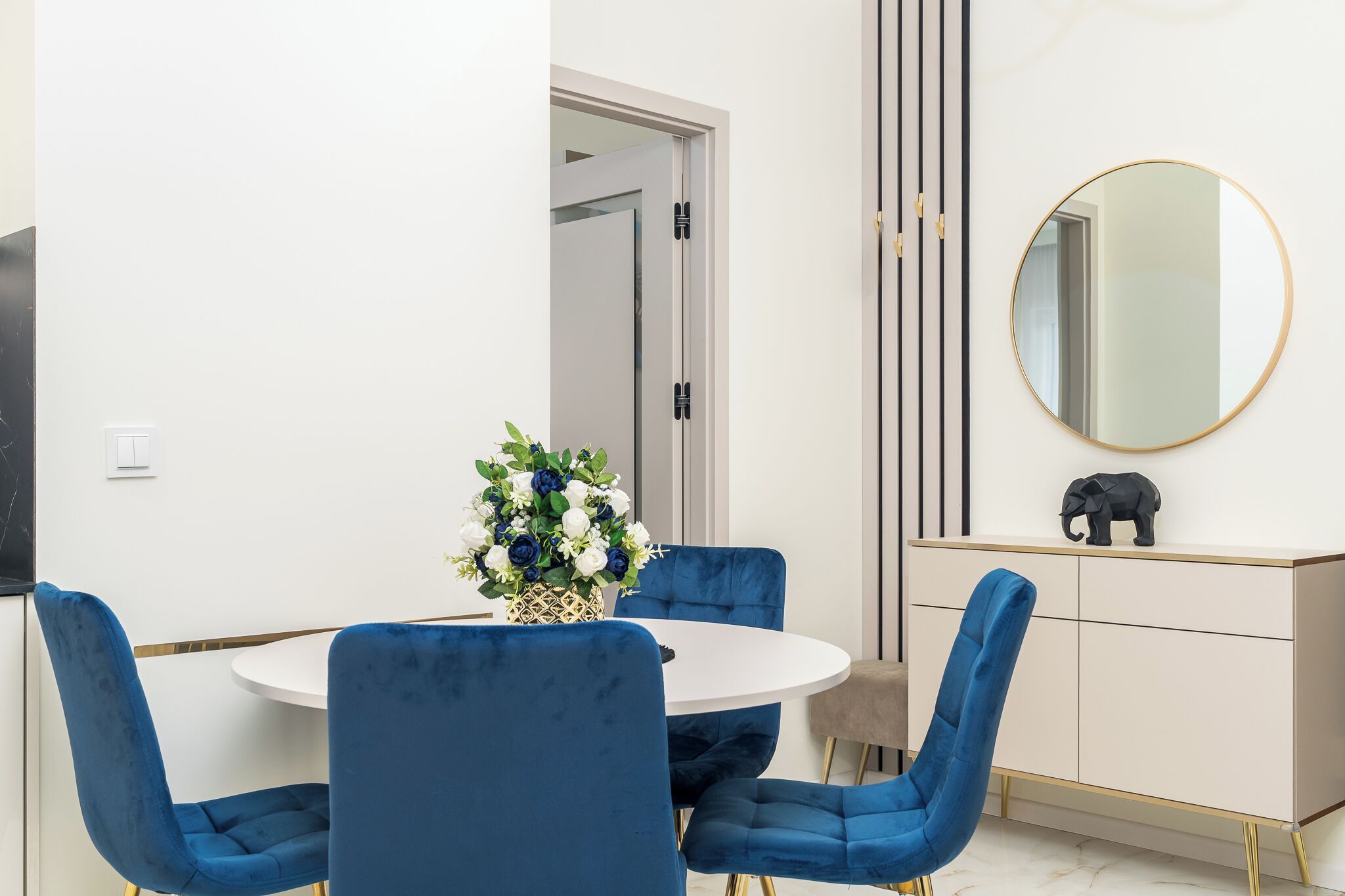 A dining area with a round table, blue chairs, a vase of flowers, a white cabinet with a gold elephant decoration, and a large round mirror on the wall.