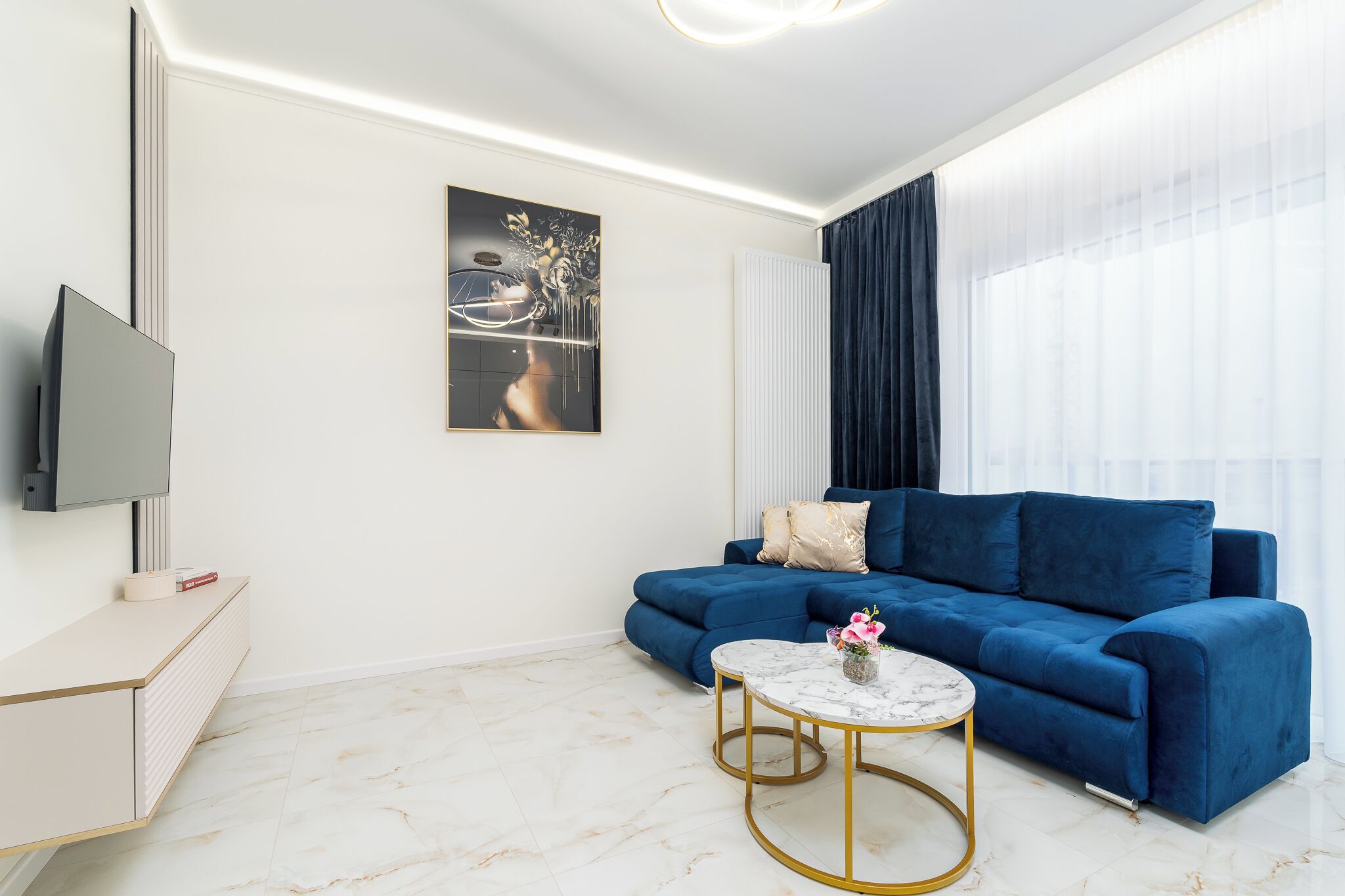 Una sala de estar moderna con un sofá azul, una mesa de centro de mármol, un televisor de pantalla plana y un ventanal con cortinas ligeras.