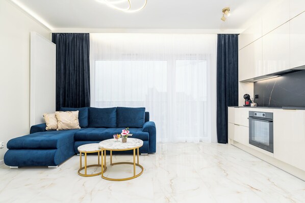 A modern living room with a blue sectional sofa, a small round table with a vase, and a kitchen area with a stove and oven. - MARGI Maris Apartament (Szczecin)
