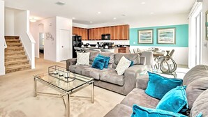 Ein modernes Wohnzimmer mit einem Glastisch, einem grauen Sofa mit blauen Kissen und einer Küche im Hintergrund.