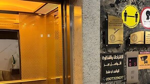 Interior de un ascensor con pared de mármol y puerta de vidrio. Hay varios letreros, incluyendo uno de prohibido fumar, otro de distanciamiento social, y uno con un número de teléfono.