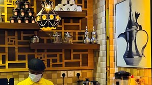 Una cocina moderna con un chef preparando comida, un electrodoméstico cilíndrico peculiar y una pared decorativa con un jarrón grande.