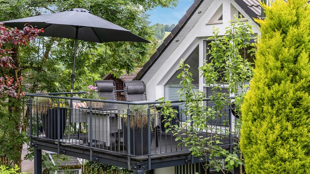 Un área de estar al aire libre techada con mesa y sillas, una sombrilla negra y vista a una casa con balcón.