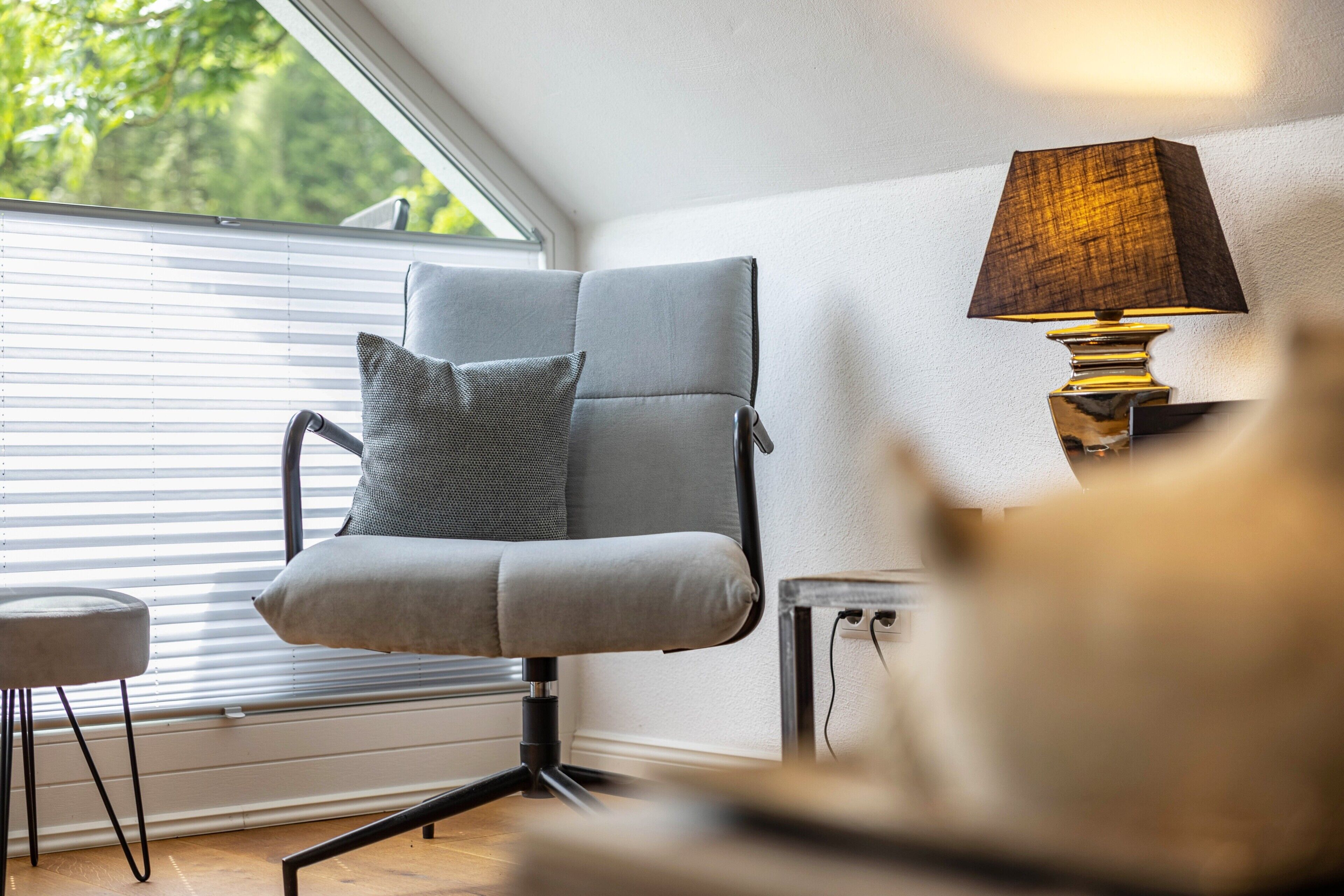 A modern chair with a cushion, a lamp, and a window with blinds.