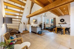 En rummelig opholdsstue med trappe, træbjælker, sofa, spisebord og en lysekrone.