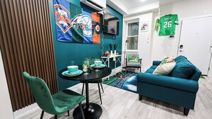 Ein modernes Wohnzimmer mit einem türkisfarbenen Sofa, einem Esstisch mit Stühlen und sportlicher Dekoration, darunter eine Flagge der Philadelphia 76ers und ein grünes Trikot mit der Nummer 26.