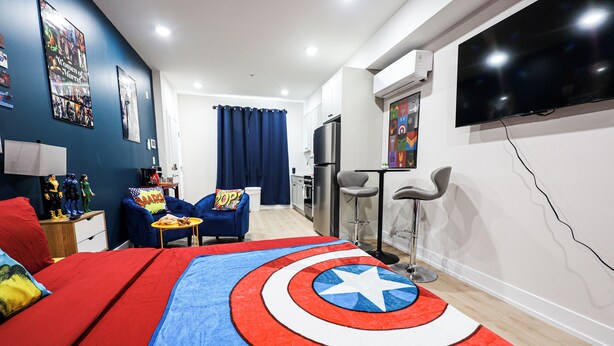 A bedroom with a Captain America themed bedspread, a TV mounted on the wall, and a kitchenette area. - Superhero Stay Marvel Fan Paradise. (Philadelphia)