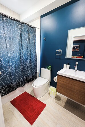 A bathroom with a blue wall, a white toilet, a white sink, and a red rug. - Superhero Stay Marvel Fan Paradise. (Philadelphia)