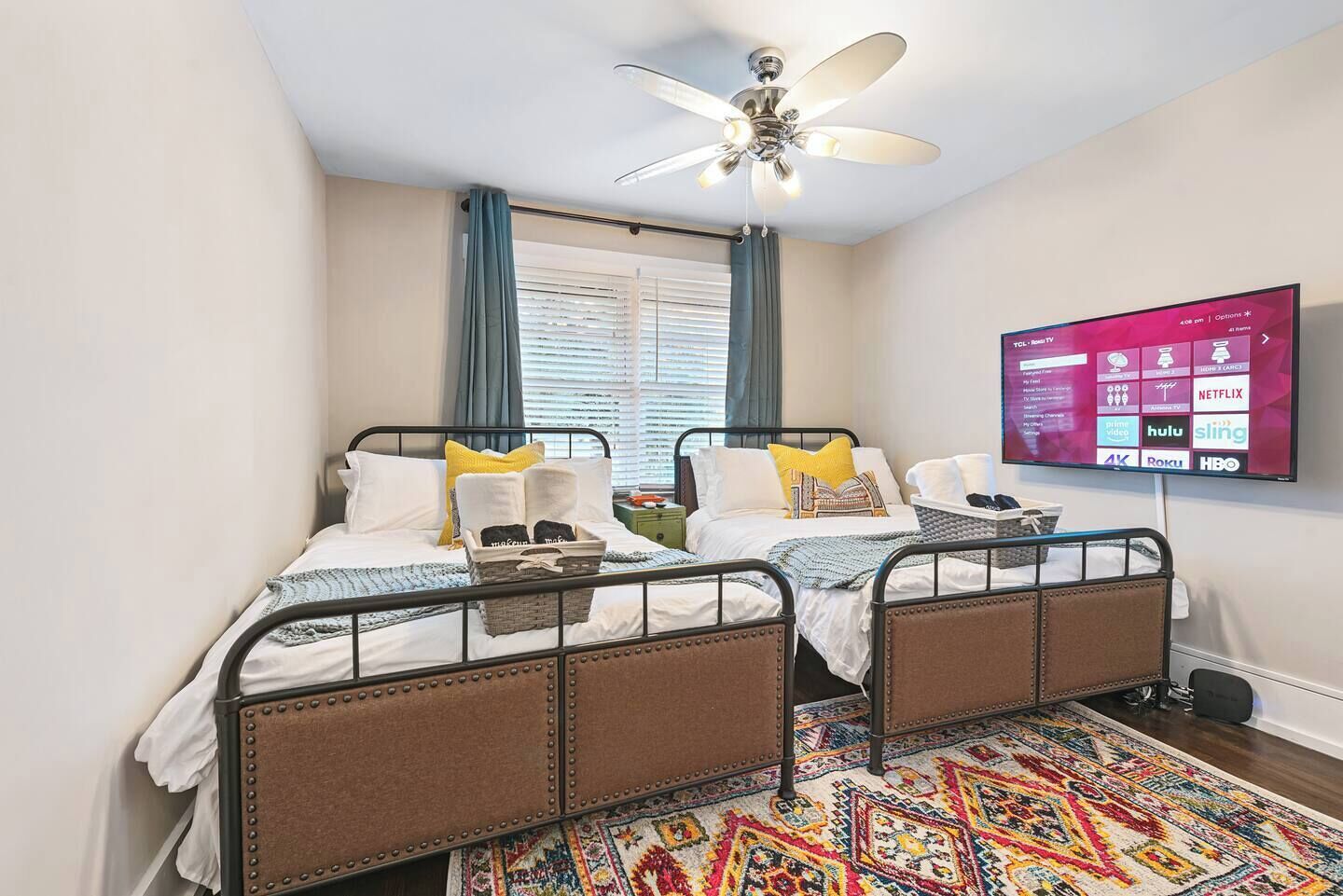 A bedroom with two beds, a ceiling fan, a TV displaying streaming services, and a colorful rug.