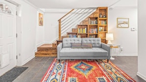 Ein Wohnzimmer mit grauem Sofa, einem bunten Teppich, einer Holztreppe und einem Bücherregal.
