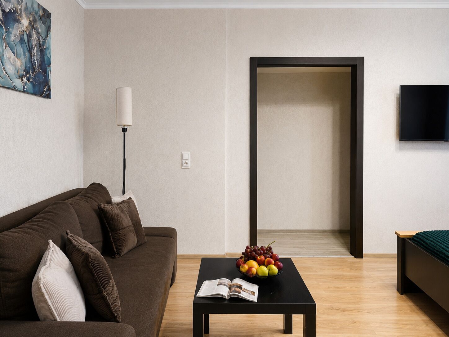 A modern living room with a brown sofa, a black coffee table with a fruit bowl and magazine, a floor lamp, and a TV on the wall.