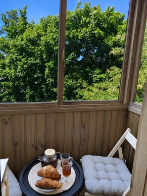 A wooden deck with a view of green trees, a round table with croissants and drinks, and a cushioned chair. - Apartment near Komarovsky Market (Minsk)