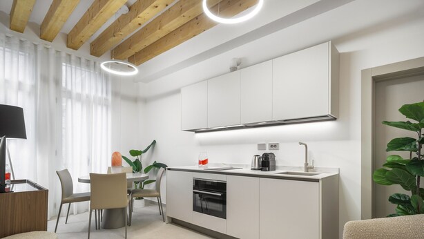 Una cocina moderna con armarios blancos, horno empotrado y cafetera. Hay un comedor con mesa y sillas, y una planta en la esquina.