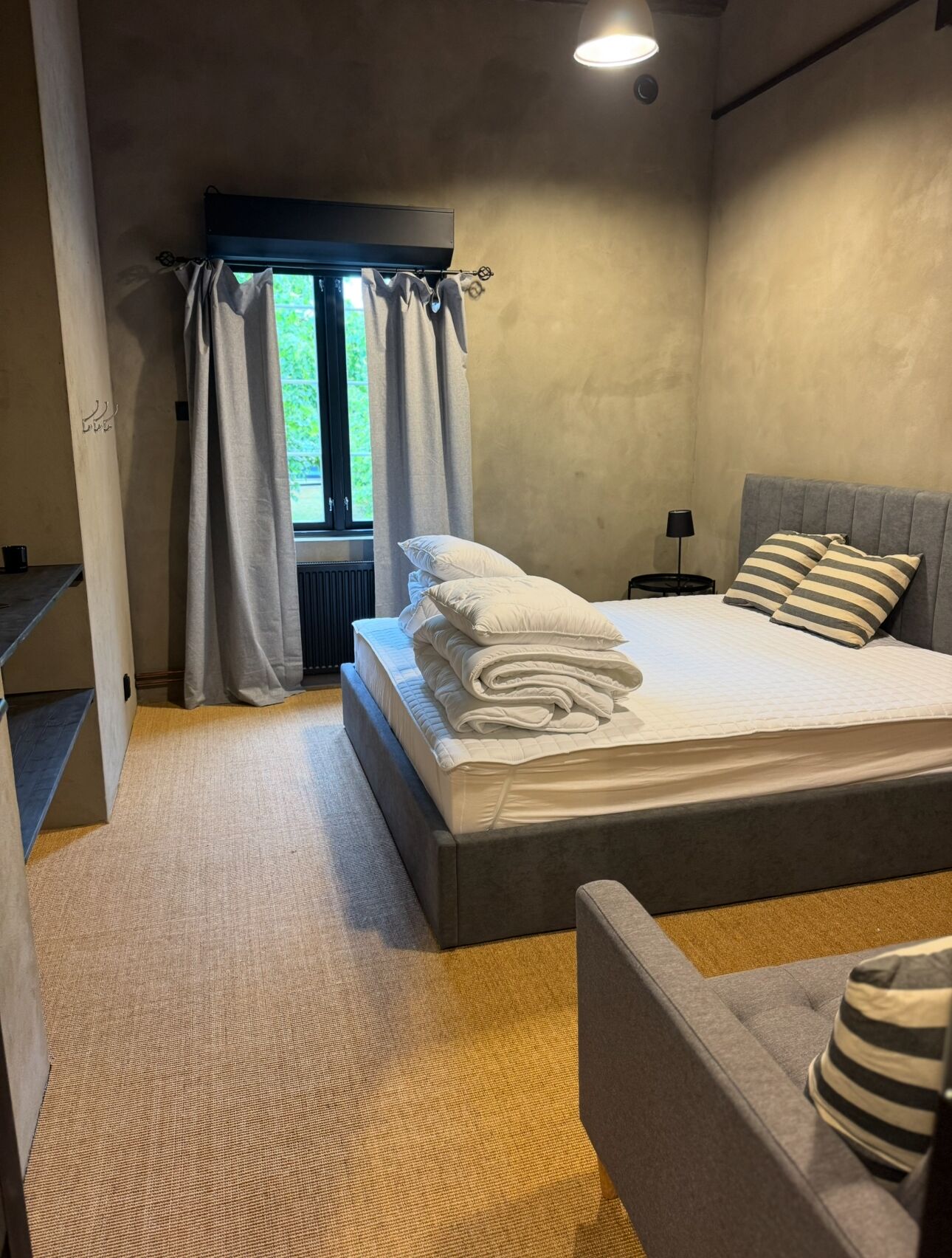 A modern bedroom with a large bed, a small bedside table, a chair, and a window with curtains.