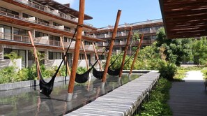 A modern hotel with hammocks over a water feature, a covered walkway, and a pool area. - Premium Condo with 2 Pools, Full Gym & Sun Decks (Tulum)