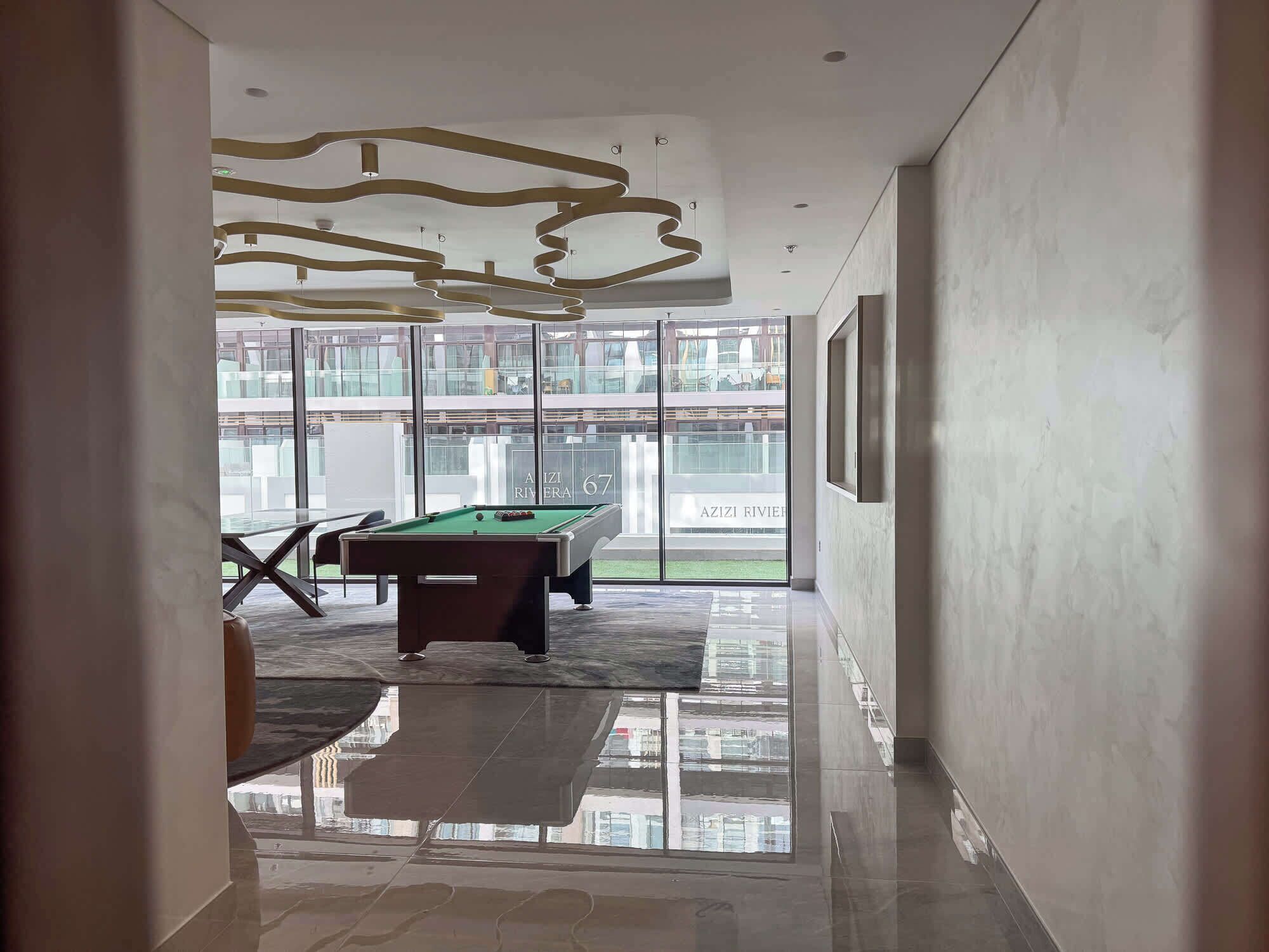 A modern interior with a pool table, a billiards cue rack, and a large window offering a view of a building with balconies.