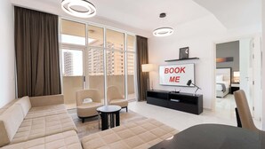 A modern living room with a sofa, chairs, a TV, and a balcony with curtains. - StayShort - 1 Bed in JVC Sleeps 4 with City Views! (Jumeirah Village Circle)