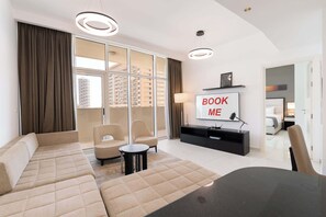A modern living room with a sofa, chairs, a TV, and a balcony with curtains. - Free housekeeping visits during your stay - StayShort - 1 Bed in JVC Sleeps 4 with City Views! (Jumeirah Village Circle)