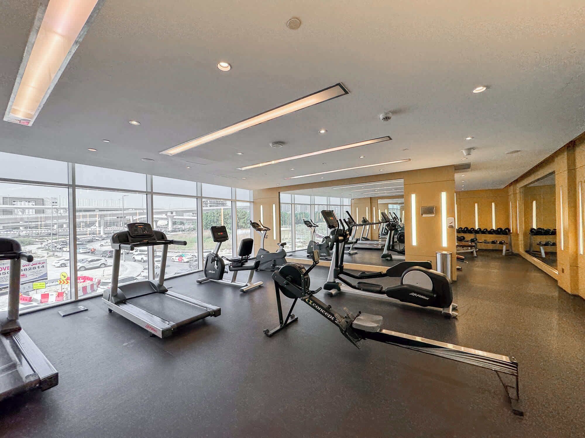 A modern gym with various cardio machines, weightlifting equipment, and large windows offering city views.