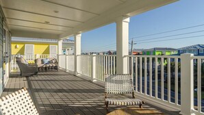 A covered porch with white railings, wicker furniture, and a view of colorful houses in the background. - Spacious Home with Private Pool Across from Ocean (Kure Beach)