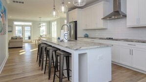 A modern kitchen with white cabinets, a large island with bar stools, stainless steel appliances, and pendant lighting. - Spacious Home with Private Pool Across from Ocean (Kure Beach)