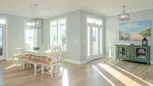 A spacious dining area with a white table and benches, large windows, and a green cabinet with a lantern. - Spacious Home with Private Pool Across from Ocean (Kure Beach)