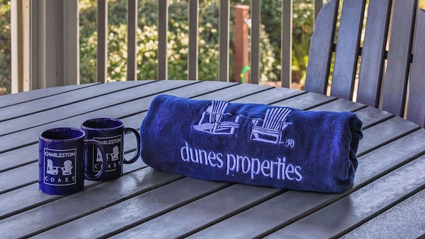 Een houten tafel met twee blauwe mokken met de tekst 'Charleston Coast' en een blauwe handdoek met 'Dunes Properties'.