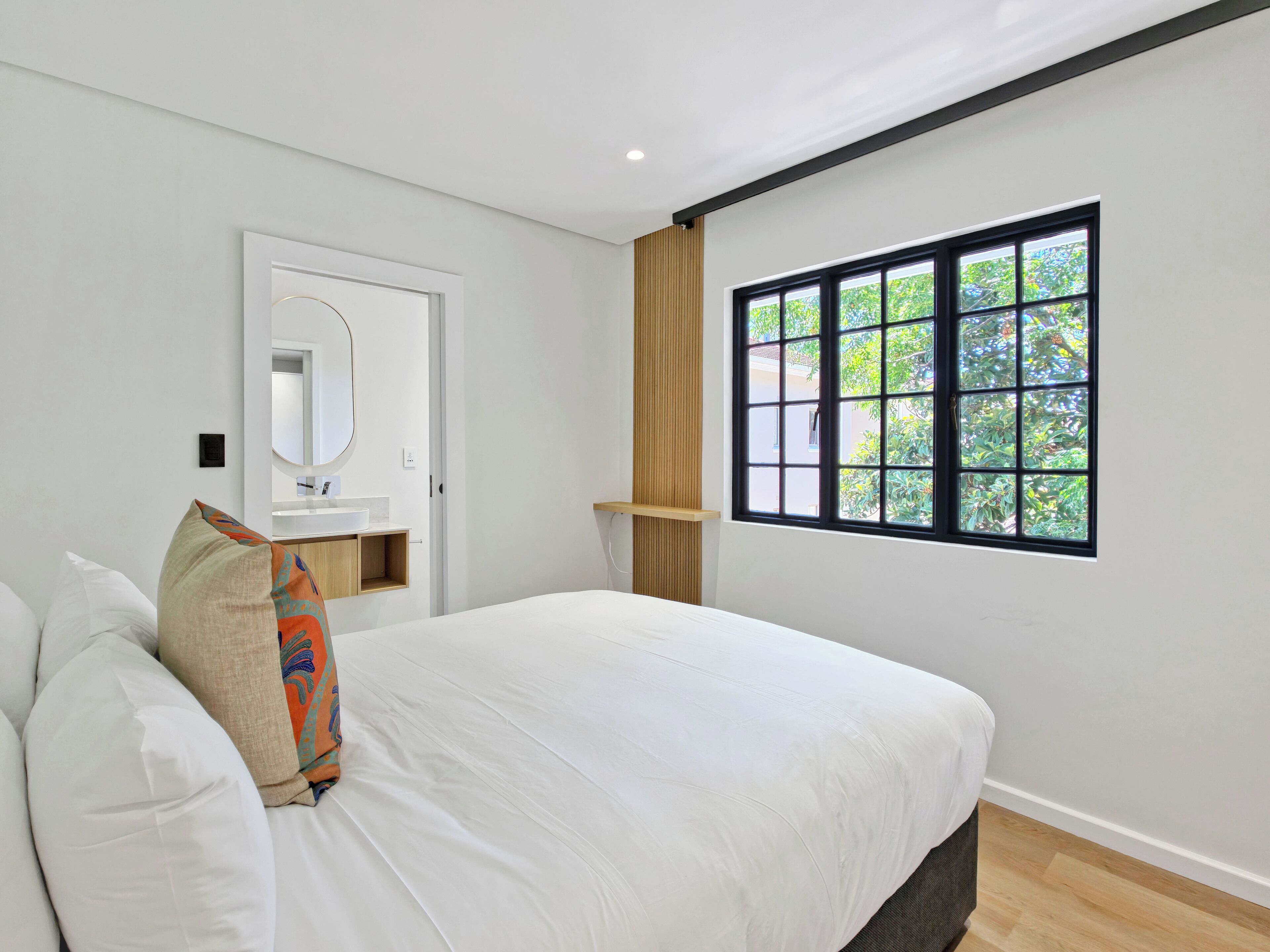 A bedroom with a large bed, a bedside table, a mirror, a window with a view of greenery, and a wooden shelf.