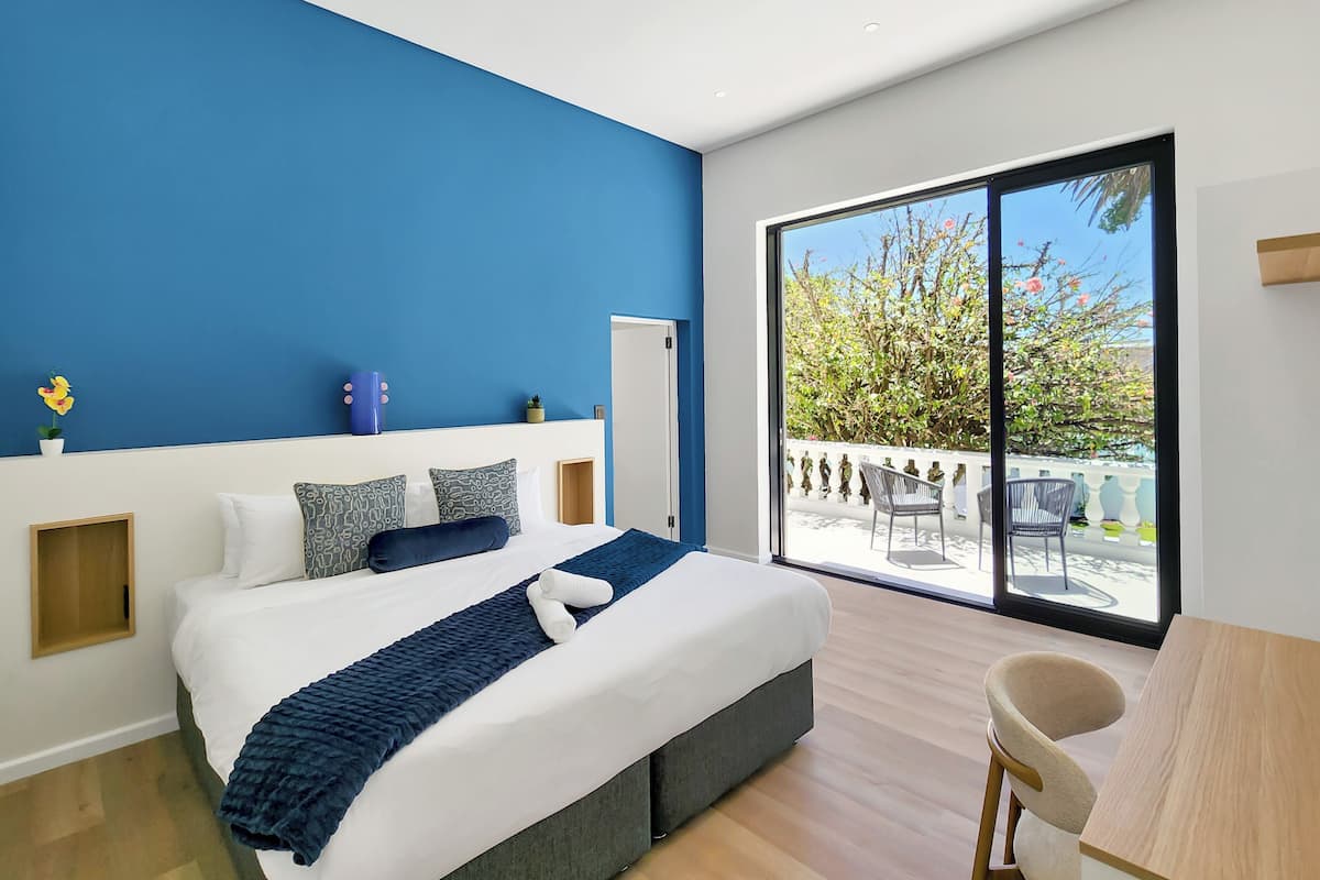 A modern bedroom with a large bed, a desk, and a balcony with a view of trees and a fence.