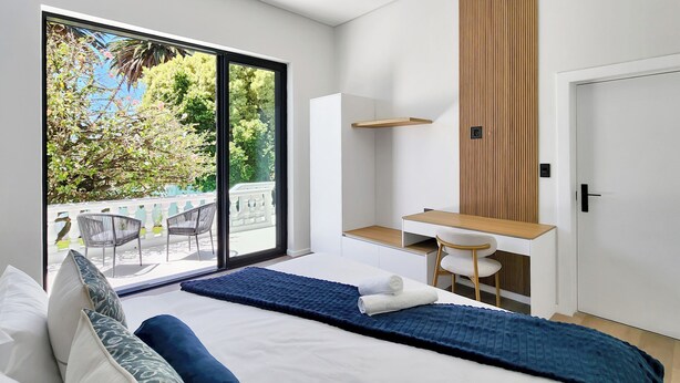 A modern hotel room with a large balcony, a desk, and a bed with blue bedding. - Palm Paradise (Cape Town)