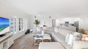 A modern living room with a large window, a flat-screen TV, a sofa, a coffee table, and a kitchen area with cabinets and appliances. - Palm Beach 41A by Liquid Life (Orange Beach)