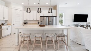 A modern kitchen with white cabinetry, stainless steel appliances, a large island with bar stools, pendant lighting, and a flat-screen TV. - Spacious Retreat Made for Memory-Making Moments w/ Pool, Spa, and Game Room (Inlet Beach)