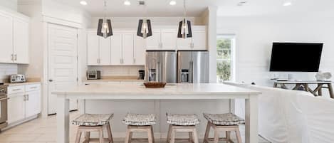 A modern kitchen with white cabinetry, stainless steel appliances, a large island with bar stools, pendant lighting, and a flat-screen TV.