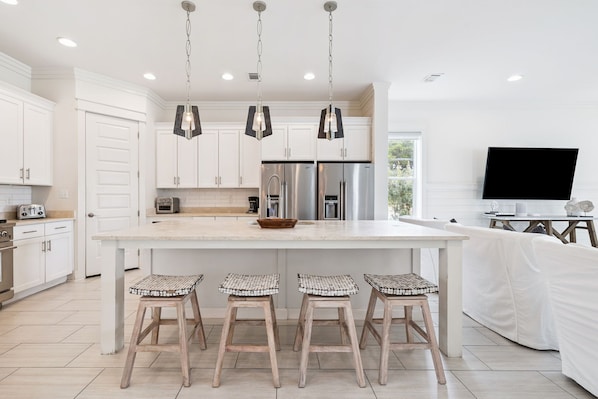 Una cocina moderna con armarios blancos, electrodomésticos de acero inoxidable, una isla grande con taburetes de bar, iluminación colgante y un televisor de pantalla plana.