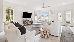 A spacious living room with white furniture, a flat-screen TV, and large windows. - Spacious Retreat Made for Memory-Making Moments w/ Pool, Spa, and Game Room (Inlet Beach)