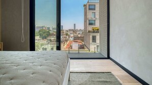 A bedroom with a large bed, a view through a sliding door to an outdoor balcony, and a cityscape in the background. - Brand New 1 BR Condo in Roma Norte - Rooftop (Mexico City)