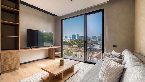 A modern living room with a large window, a flat-screen TV, a wooden coffee table, and a sofa with textured cushions. - Brand New 1 BR Condo in Roma Norte - Rooftop (Mexico City)