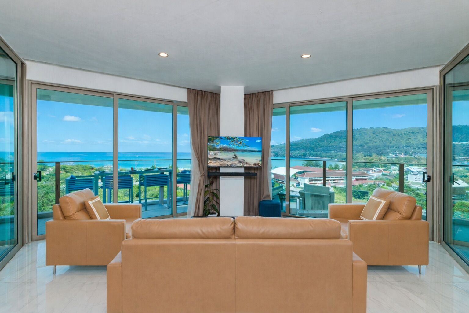 A spacious living room with a large sofa, two armchairs, and a flat-screen TV. The room features floor-to-ceiling glass doors offering a view of the ocean and a mountainous landscape.