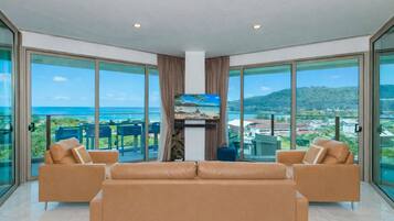 A spacious living room with a large sofa, two armchairs, and a flat-screen TV. The room features floor-to-ceiling glass doors offering a view of the ocean and a mountainous landscape.