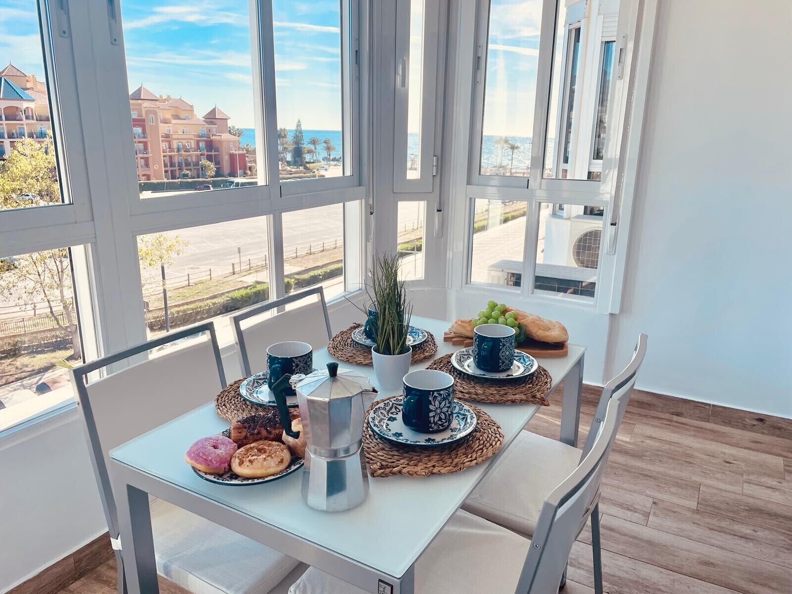 A dining table with a coffee maker, cups, and pastries in a room with large windows.