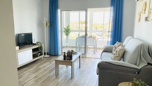 Una sala de estar moderna con un sofá gris, una mesa de centro, un televisor sobre un mueble bajo y un gran ventanal corredero que da a un espacio exterior.