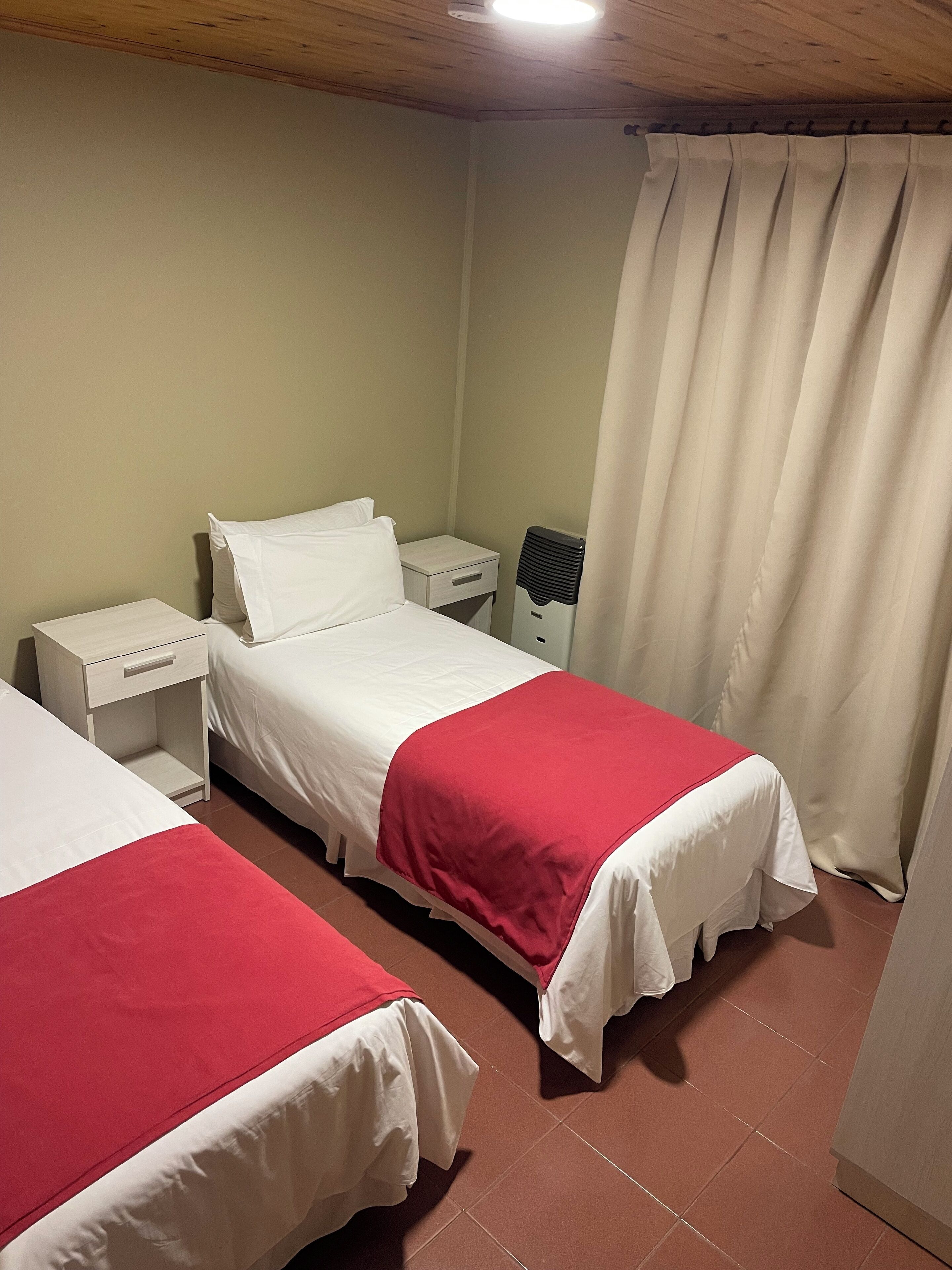 A hotel room with two single beds, a bedside table with a lamp, a heater, and a window with curtains.