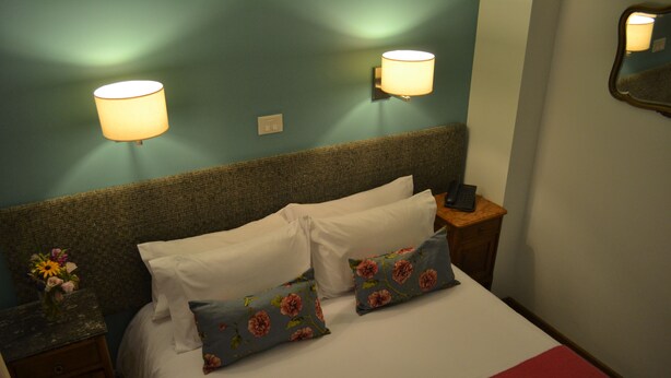 A neatly made bed with white linens and floral-patterned pillows, a bedside table with a vase of flowers, and two wall-mounted lamps. - Individual room in the Heart of the town (El Calafate)