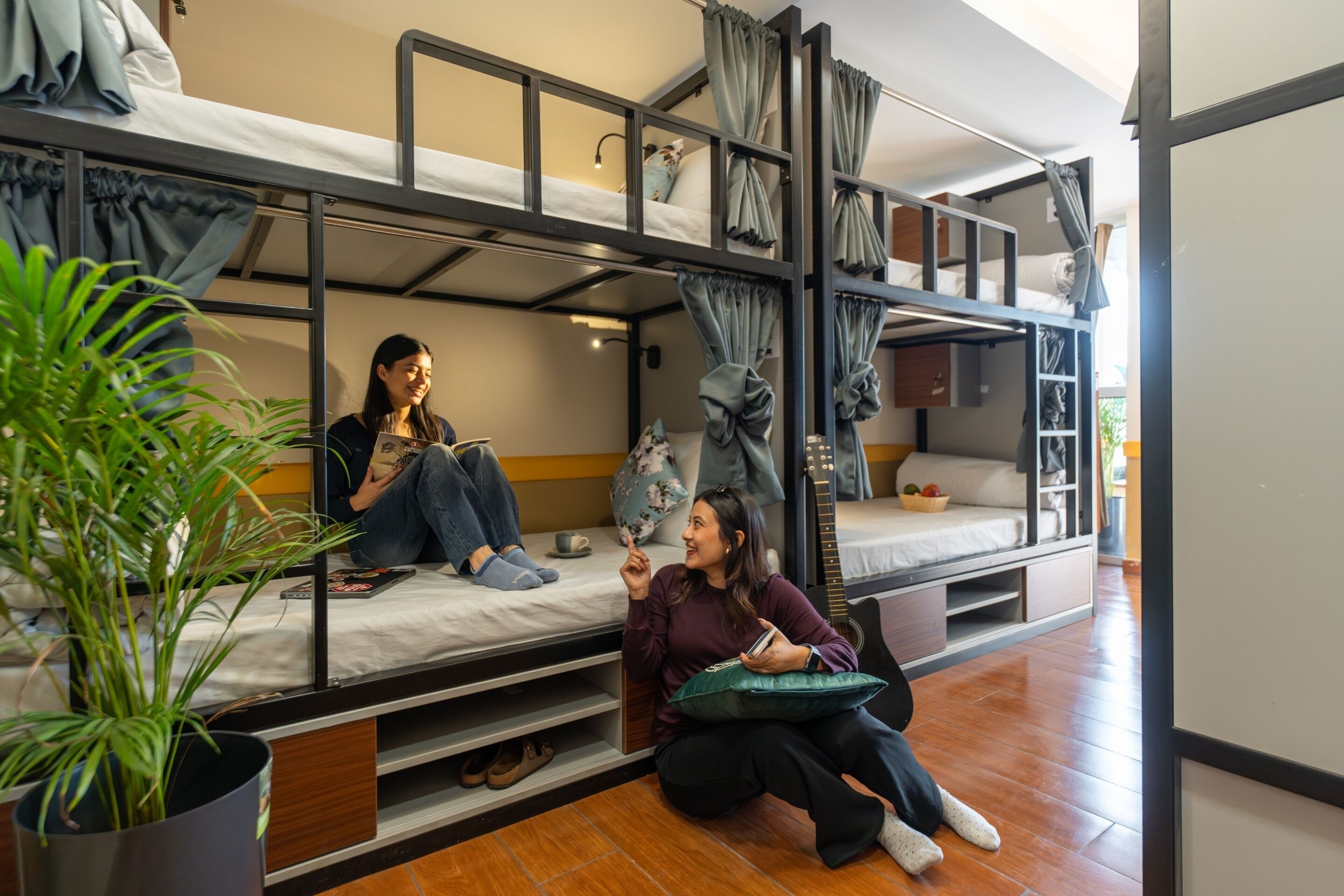 Une chambre de dortoir avec des lits superposés, une plante en pot et une personne assise par terre.