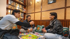 Eine Gruppe von Personen spielt auf einem Teppich Monopoly mit einem Spielbrett, Würfeln und Spielfiguren.