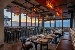 Un área de comedor al aire libre con mesas preparadas para dos, ventanas amplias que ofrecen vistas a las montañas y lámparas colgantes de estilo farol.