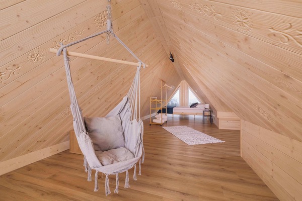 A wooden attic corner featuring a hammock chair and bookshelves. The warm wood creates a cabin-style retreat atmosphere.