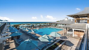 Zona de piscina en un resort con múltiples tumbonas y sombrillas, vista despejada del océano y una zona de estar cubierta.