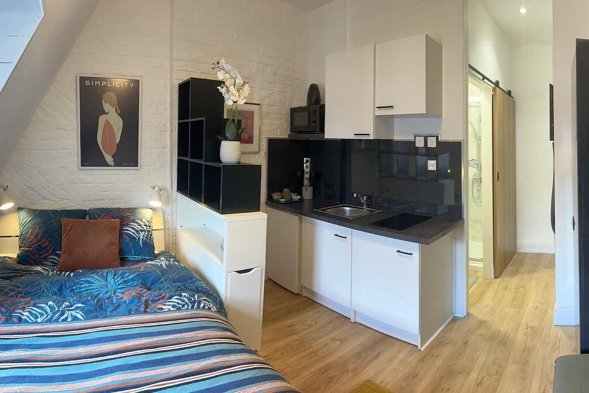 Een compact, modern appartement met een kitchenette, een bed met gestreept beddegoed en een groot raam dat voor natuurlijk licht zorgt.